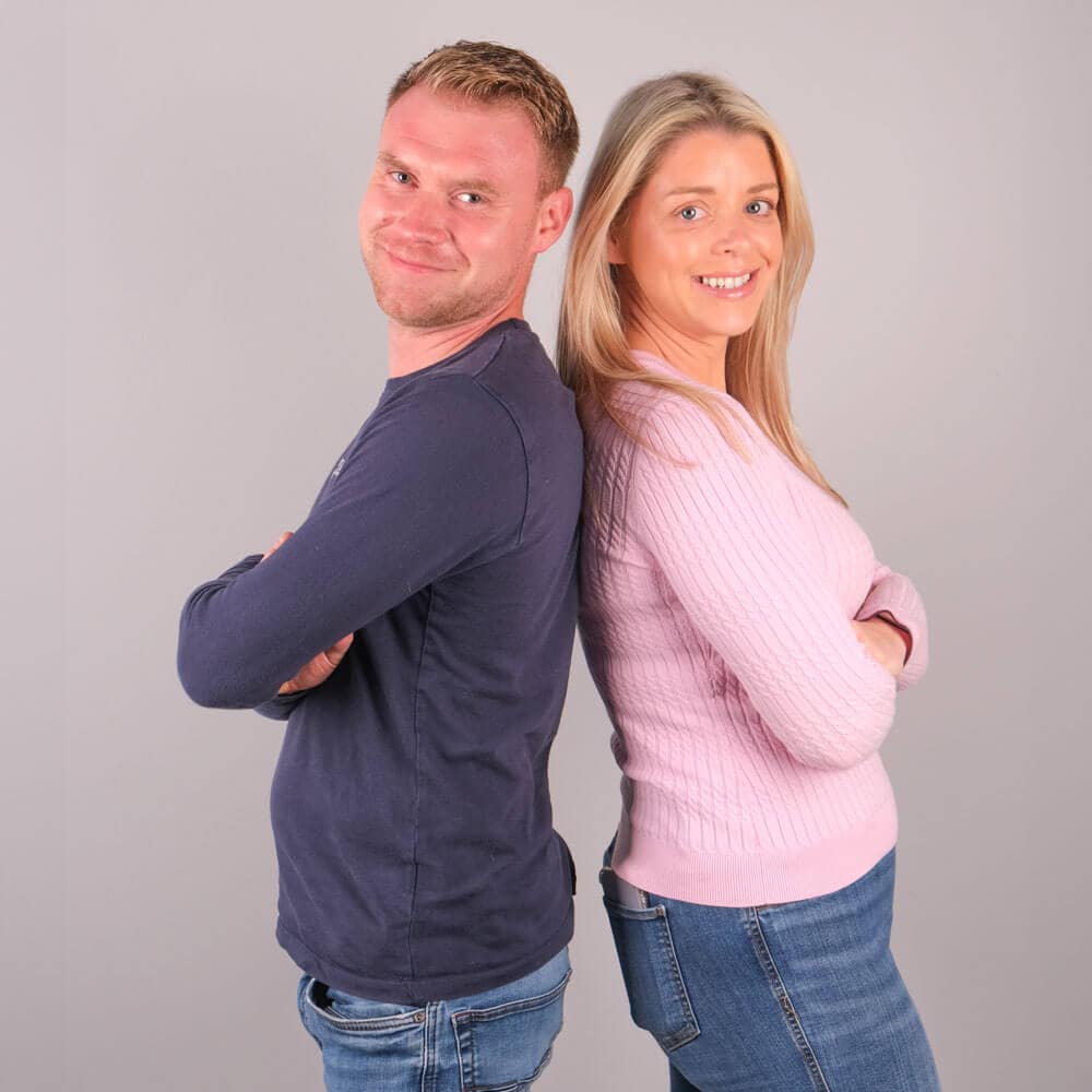 A man and a woman standing back-to-back against a plain gray background. Both have their arms crossed and are smiling. The man is wearing a dark long-sleeve shirt and jeans, while the woman is wearing a light pink sweater and jeans.