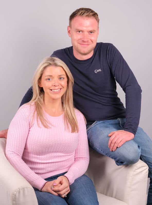 A woman with long blonde hair wearing a pink sweater is sitting on a white armchair. A man with short hair wearing a black long-sleeve shirt and blue jeans is standing beside her, resting one hand on the chair. They are both smiling against a plain gray background.