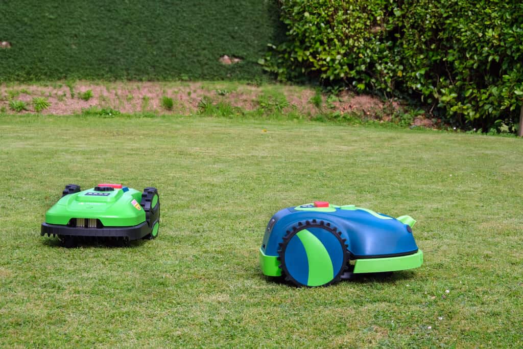 Two robotic lawn mowers, one light green and one dark green, are working on a neatly manicured lawn. One of the mowers is in the background and the second one is closer to the camera. A lush green hedge line forms the backdrop.