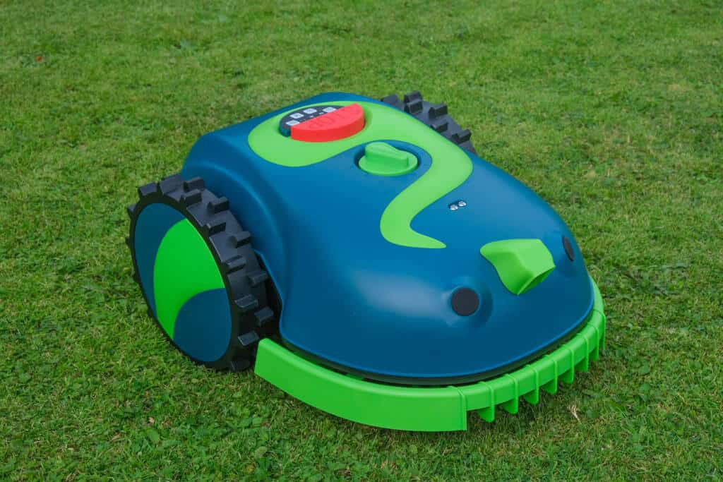 A dark green and light green robotic lawnmower sits on a grassy lawn. The robot has large, black, rugged wheels on either side and a bright red emergency stop button on top. It features green decorative swirls on the top surface, giving it a playful appearance.