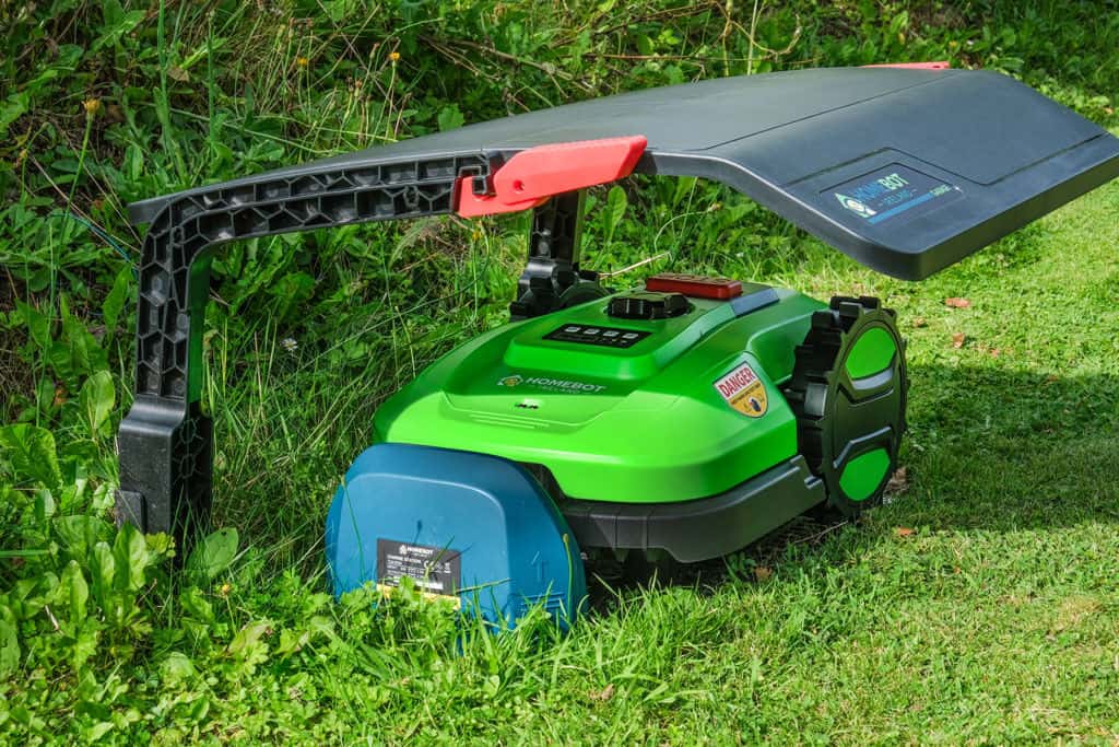 A green robotic lawn mower with a black and red cover is parked on the grass, next to bushes and plants. The mower has a dark green front and various controls and stickers on its top.