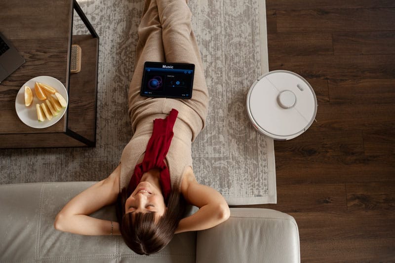 A woman lies on a rug with a tablet on her stomach, listening to music. Nearby, a HomeBot robot vacuum and mop diligently glides across the wooden floor. A plate with orange slices sits on an adjacent table, capturing the essence of a cozy home. She appears relaxed, resting her arms above her head.