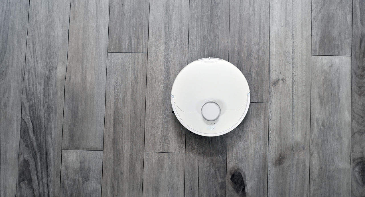 A white circular robotic vacuum cleaner is positioned on a wooden floor with grey planks in a spacious room. The floor is made up of long, narrow, and evenly spaced wood panels. The vacuum cleaner features minimalistic design elements and a central sensor.