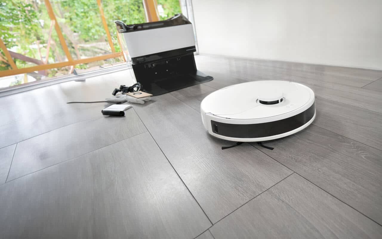 A white robot vacuum cleaner moves across a sleek, light grey hardwood floor in a modern room with large windows. Nearby, a charging station and some cleaning accessories are placed on the floor. Trees and greenery are visible through the windows.