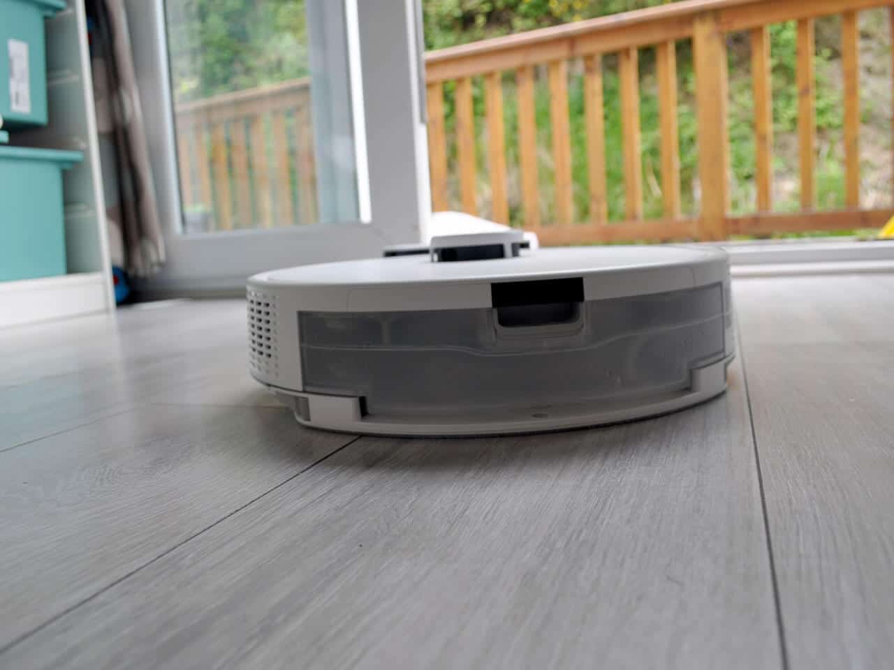 A sleek, white HomeBot Ireland robot vacuum and mop is operating on a hardwood floor near an open sliding glass door. Outside, there is a wooden railing, and the area appears to be part of a home with a garden or yard visible in the background.