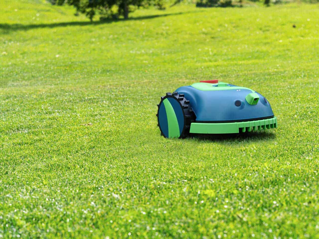 The HomeBot Ireland Buddy, a robotic lawn mower, is autonomously cutting the grass on a well-manicured green lawn under clear skies. The mostly green device with black wheels navigates the spacious outdoor area efficiently. In the background, trees and additional grassy expanses add to the serene setting.