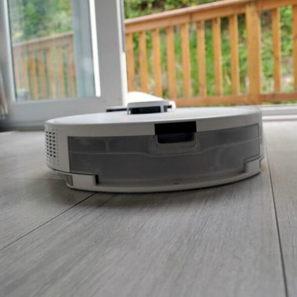 A close-up of the Spritz Robot Vacuum and Mop on light-colored wooden flooring shows it in action near a sliding glass door, with a wooden deck visible in the background. The vacuum appears to effortlessly vacuum and mop spotless cleanliness onto your floors.