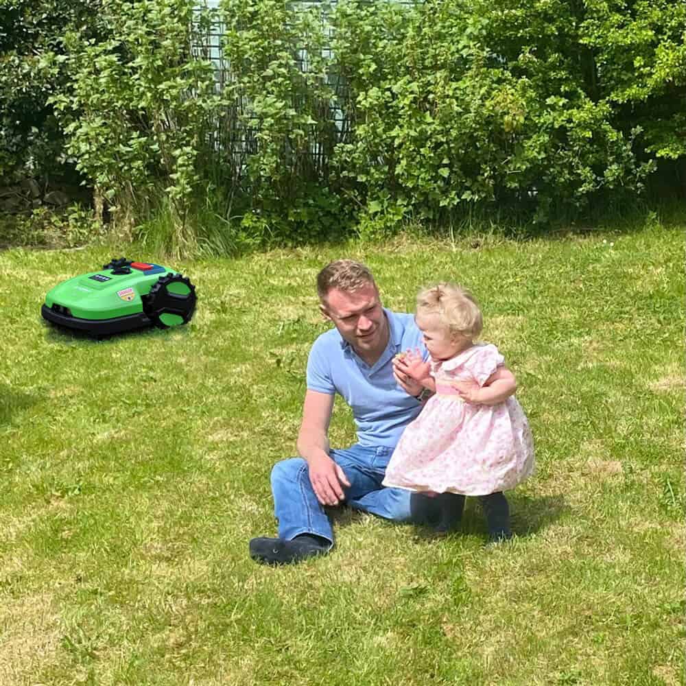 A man and a young girl are sitting on the grass in a garden. The man is sitting cross-legged while the girl, dressed in a pink dress, stands near him. In the background, there is a green robotic lawn mower from HomeBot Ireland on the grass. Bushes and trees are visible in the background.