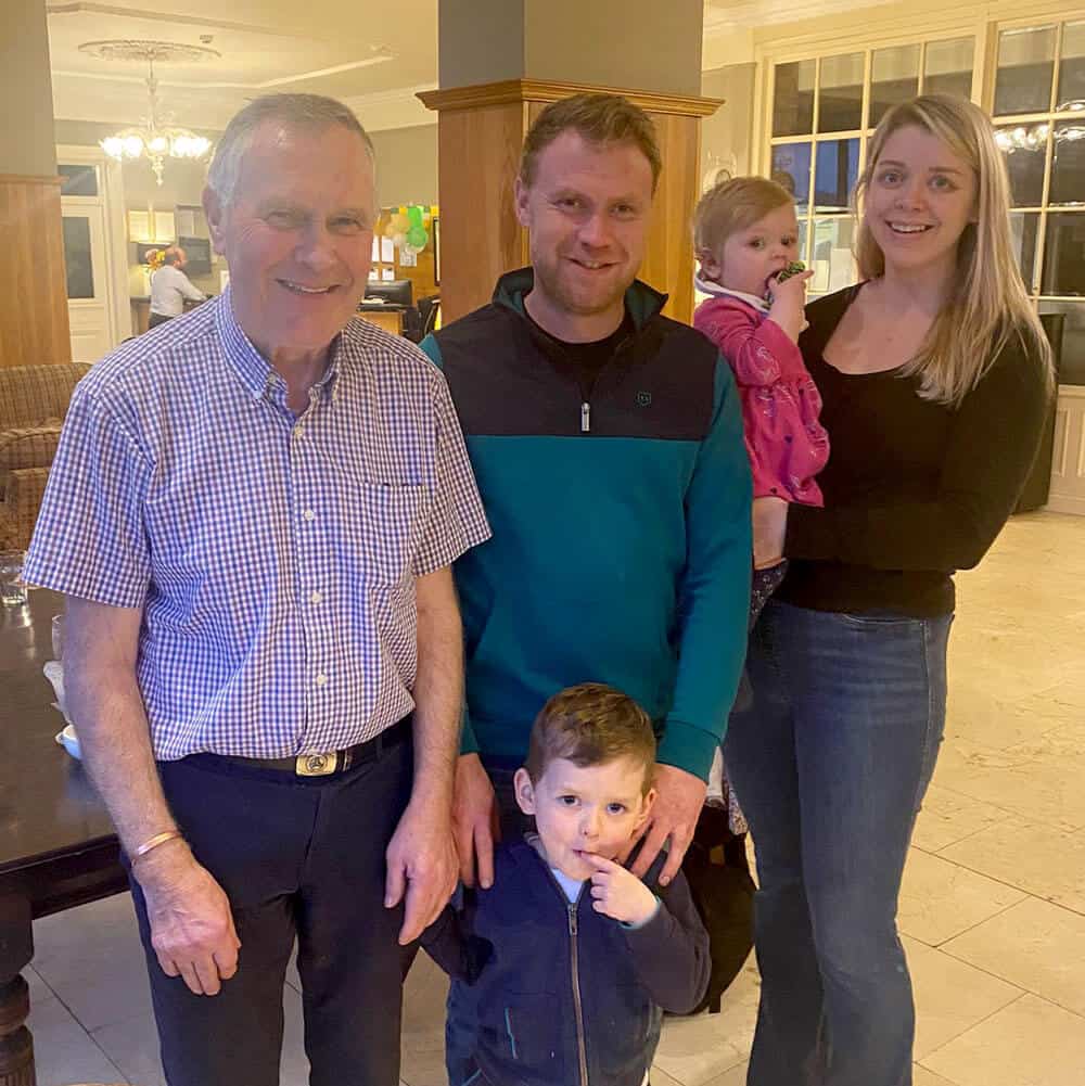 A group of five people, including an older man, a younger man, a woman holding a toddler, and a young boy stand closely together inside a warmly lit room. The older man is dressed in a checkered shirt, while the others wear casual clothing. They are part of their family business, HomeBot Ireland.