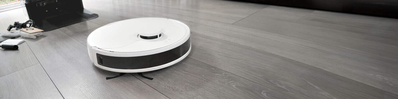 A white HomeBot Ireland robotic vacuum and mop is moving across a clean, light grey hardwood floor. In the background, there are various objects, including a charging station and a few small items on the floor.