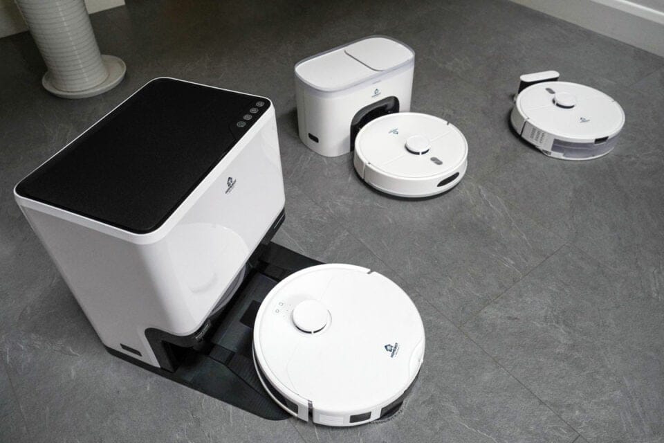 A group of white HomeBot robot vacuums and mops sit on a gray tiled floor. From left to right - Glider, Dottie and Spritz with their charging stations.