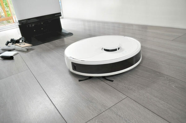 A sleek white Glider Robot Vacuum and Mop glides across a light gray wooden floor, effortlessly picking up dust. In the background, there is a black charging dock with some cables and a remote control placed nearby. The scene appears to be inside a modern, minimalist home.