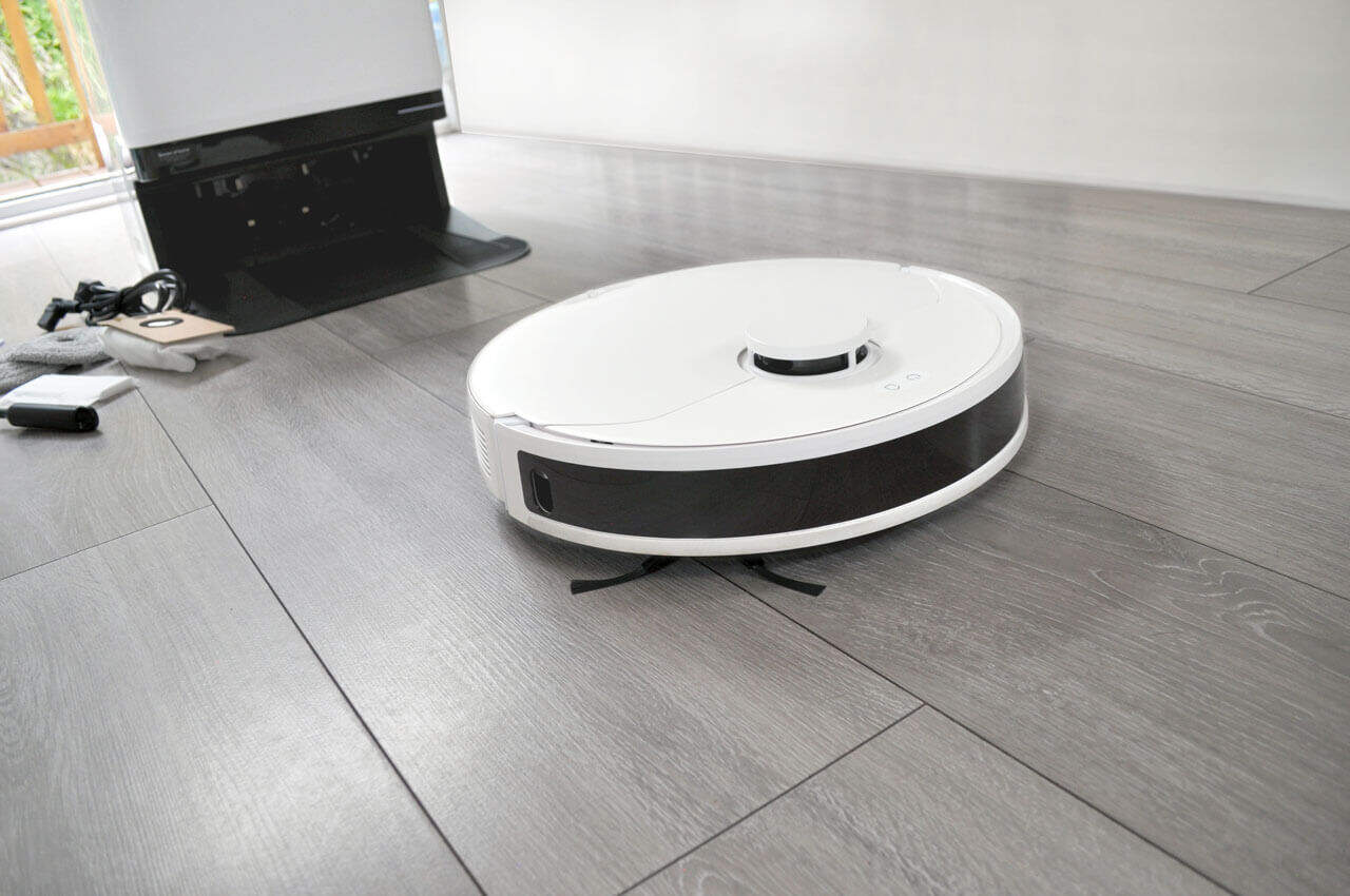 A white robotic vacuum cleaner is operating on a sleek, light gray wooden floor. In the background, there is a charging dock with some related equipment and cables nearby. The scene is well-lit, and the floor looks clean.