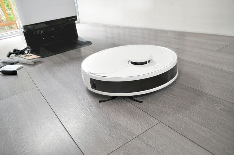 A white HomeBot Ireland robot vacuum and mop is seen on a light gray wooden floor, near a charging dock with cables and a remote control beside it. The room has a minimalistic design with natural light streaming in from the left side.